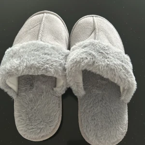 Grå tofflor med fuskpäls - Mysiga grå tofflor med mjuk fuskpäls runt kanten och insidan. Perfekta för att hålla fötterna varma under kalla dagar. Sulan är ribbad för extra grepp.