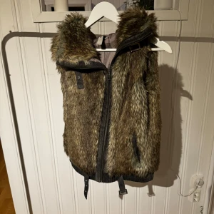 Brun pälsväst från Mind - Säljer en snygg brun pälsväst från Mind med dragkedja och huva. Västen har en cool look med pälsdetaljer och är perfekt för att hålla sig varm och stilren. Passar bra till både vardag och speciella tillfällen.