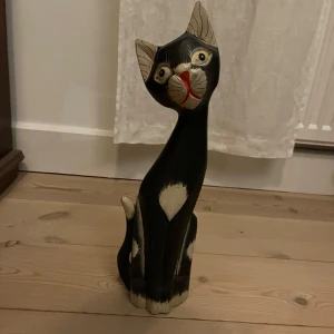 Träskulptur av katt - Charmig träskulptur föreställande en svart katt med vita detaljer. Katten har en elegant och stiliserad form med ett uttrycksfullt ansikte. Perfekt som dekoration i hemmet för kattälskare. Höjd 41 cm. Har totalt 4 st i olika storlekar 