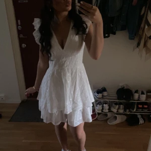 Vit klänning med volangdetaljer - Säljer en vacker vit klänning med volangdetaljer och v-ringning. Klänningen har ett fint blommigt mönster. Jag har använt den en gång på en avslutning för något år sedan och har ingen användning av den längre. Den har en dragkedja i ryggen för enkel påklädning.