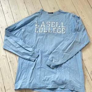 Ljusblå långärmad tröja med tryck - Säljer en ljusblå långärmad tröja med texten 'Lasell College' tryckt på framsidan. Perfekt för en avslappnad stil. Tröjan har en normal passform och är tillverkad i ett mjukt material.