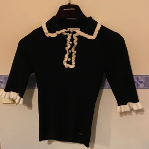 Svart tröja med vita detaljer från Holly & Whyte - Snygg svart ”Chanel-inspirerad” tröja från Holly & Whyte med vita volangdetaljer vid krage och ärmslut. Tröjan har trekvartsärmar och knappar framtill. Perfekt för en stilren look. Oanvänd 