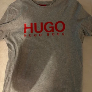 Grå t-shirt från Hugo Boss - Snygg grå t-shirt från Hugo Boss med röd logga på bröstet. Perfekt för en stilren och avslappnad look. Kortärmad och tillverkad i mjukt material för extra komfort.