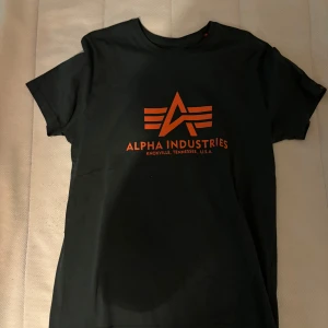 Svart t-shirt från Alpha Industries - Snygg svart t-shirt från Alpha Industries med orange logotyp och text på framsidan. Perfekt för en avslappnad stil.