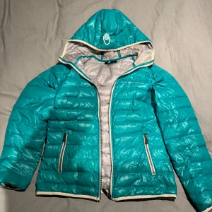 Turkos dunjacka från Sail Racing - Säljer en snygg turkos dunjacka från Sail Racing. Jackan har en huva och dragkedja framtill samt två praktiska fickor med dragkedjor. Perfekt för kyliga dagar med sin quiltade design och grå insida. Lite skitig runt toppen av dragkedjan annars inget skit eller skador. 