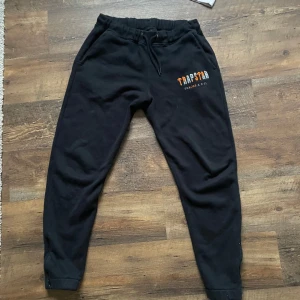 Svarta mjukisbyxor från Trapstar - Säljer ett par svarta mjukisbyxor från Trapstar med elastisk midja och muddar vid bensluten. Byxorna har en tryckt logga i orange och vit på ena benet. Perfekta för en avslappnad stil. (Bara äkta kläder)