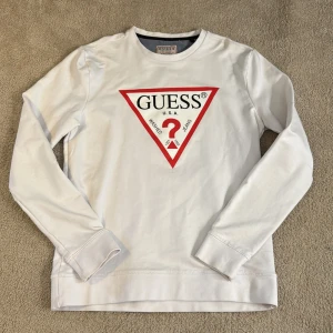 Vit långärmad tröja från Guess - Säljer en vit långärmad tröja från Guess med det ikoniska röda triangellogotypen på framsidan. Tröjan har en klassisk rund hals och är perfekt för en stilren look. Säljer pga att den inte används💗