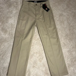 Dickies - Snygga beige byxor från Dickies med en klassisk design. Byxorna har en rak passform och är tillverkade i ett slitstarkt material. Perfekta för en avslappnad stil. De har en knapp och dragkedja framtill samt en bakficka med knapp.