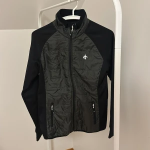 Svart sportjacka  - Snygg svart sportjacka med dragkedja framtill och två praktiska fickor. Jackan har en stilren design med långärmade ärmar och en hög krage. Perfekt för utomhusaktiviteter och träning.