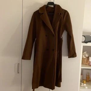 Brun kappa med bälte - Snygg brun kappa med knappar och bälte i midjan. Perfekt för höst och vinter. Långärmad design med klassisk krage.