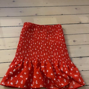 Röd blommig kjol - Säljer en söt röd kjol med vita blommor. Kjolen har en volangdetalj längst ner och är perfekt för sommarens varma dagar. Den elastiska midjan ger en bekväm passform. Använd fåtal gånger. Är omsydd från storlek m men är nu i storlek s. Väldigt strechig så passar storlek xs-m. Pris kan diskuteras!🥰