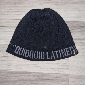 Svart mössa med text - Snygg svart mössa från Arka med texten 'QUIDQUID LATINE' i grått. Perfekt för att hålla värmen under kyliga dagar. Mössan har en enkel och stilren design som passar till det mesta.