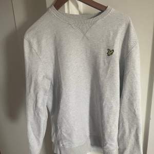 Säljer en stilren och bekväm grå tröja från Lyle & Scott. Den har en klassisk design med ett litet broderat emblem på bröstet. Perfekt för både vardag och avslappnade tillfällen.