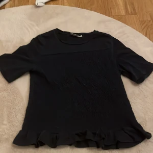 Svart topp från Gina Tricot - Säljer en snygg svart topp från Gina Tricot. Den har korta ärmar och en fin volangdetalj vid nederkanten. Perfekt för både vardag och fest!