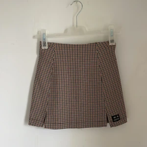 Rutig kjol från Urban Outfitters - Säljer en superfin rutig kjol från Urban Outfitters i storlek XS. Kjolen har ett klassiskt rutmönster i svart, vitt och brunt, vilket ger den en tidlös look. Den är kort och har en liten slits framtill som ger en extra touch. Perfekt för hösten.