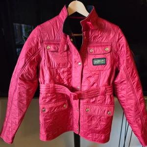Röd quiltad jacka från Barbour - Säljer en snygg röd quiltad jacka från Barbour International. Jackan har en cool röd färg med knappar och dragkedja framtill, samt ett bälte i midjan för en perfekt passform. Den har en mjuk fleece insida för extra komfort. Perfekt för vår och höst.