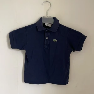 Mörkblå pikétröja från Lacoste - BARNTRÖJA‼️Säljer en snygg mörkblå pikétröja från Lacoste. Den har en klassisk krage med två knappar och det ikoniska krokodilmärket på bröstet. Perfekt för en stilren och avslappnad look!