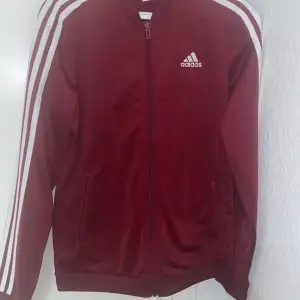 Säljer en snygg vinröd Adidas jacka med vita ränder längs ärmarna. Jackan har dragkedja framtill och är perfekt för träning eller vardagsbruk. Den är i bra skick och redo för nya äventyr!