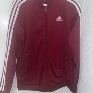 Vinröd jacka från Adidas - Säljer en snygg vinröd Adidas jacka med vita ränder längs ärmarna. Jackan har dragkedja framtill och är perfekt för träning eller vardagsbruk. Den är i bra skick och redo för nya äventyr!