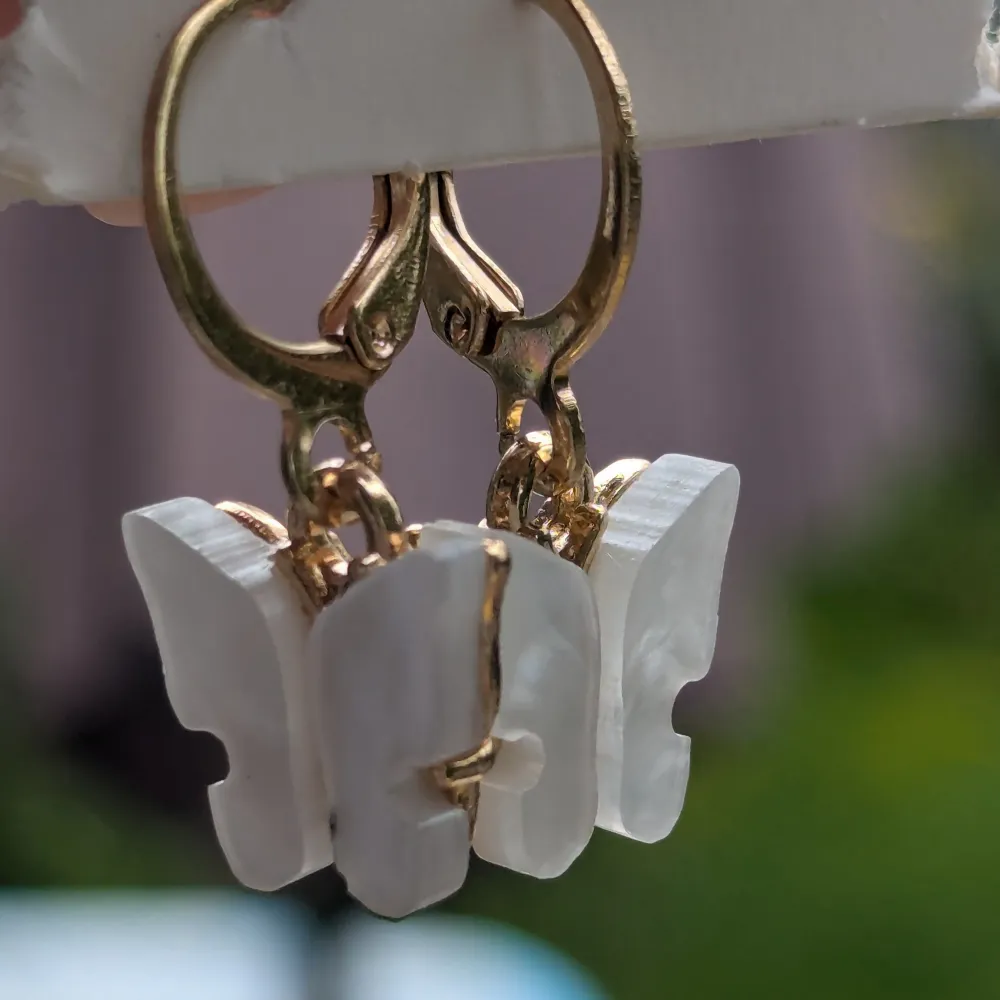 Superfina örhängen med vita fjärilar och guldfärgade detaljer. De är formade som små fjärilar och hänger på en guldfärgad ring. Perfekta för att ge en söt och elegant touch till vilken outfit som helst!. Asusteet.
