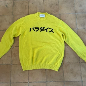 Gul stickad tröja från Axel Arigato - Säljer en cool gul stickad tröja från Axel Arigato med japansk text 'Paradise' på framsidan och ett stort yin-yang-märke på baksidan. Perfekt för att sticka ut i mängden! Tröjan har långa ärmar och en bekväm passform. Passar perfekt för höst och vår.