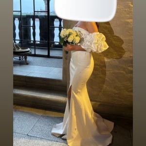 Vit axelbandslös klänning med volangärmar - Säljer en elegant vit axelbandslös klänning med puffiga volangärmar och en stilren slits. Perfekt för bröllop eller festliga tillfällen. Klänningen har en tight passform som framhäver figuren och små pärldetaljer som ger en extra touch av lyx.