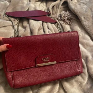 Vinröd axelväska från Guess - Säljer en snygg vinröd axelväska från Guess i skinn. Väskan har en kedjerem och ett stilrent metallspänne framtill. Perfekt för både vardag och fest! 👜
