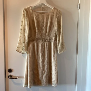 Beige klänning med paljettdetaljer - Säljer en elegant beige klänning med vackra paljettdetaljer. Klänningen har trekvartsärmar och en normal passform, perfekt för festliga tillfällen. Materialet känns lyxigt och ger en fin struktur. Den är i mycket bra skick och redo för nästa ägare!