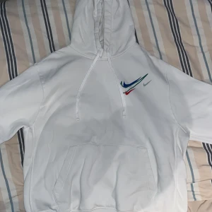 Vit hoodie från Nike - Säljer en snygg vit hoodie från Nike i storlek L. Den har en klassisk design med en färgglad Nike-logga i blått, grönt och rött på bröstet. Perfekt för både vardag och träning. Hoodien har en stor ficka framtill och justerbar huva.