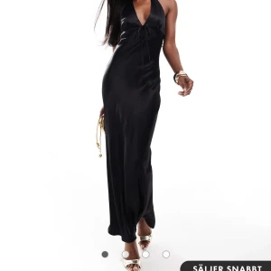 Svart långklänning i satin - Elegant svart långklänning i satin med öppen rygg och söt knytning framtill. Perfekt för festliga tillfällen med sin glänsande yta och smickrande passform. Klänningen har en djup urringning och är ärmlös, vilket ger en sofistikerad look. Den är helt oanvänd med prislapp kvar!