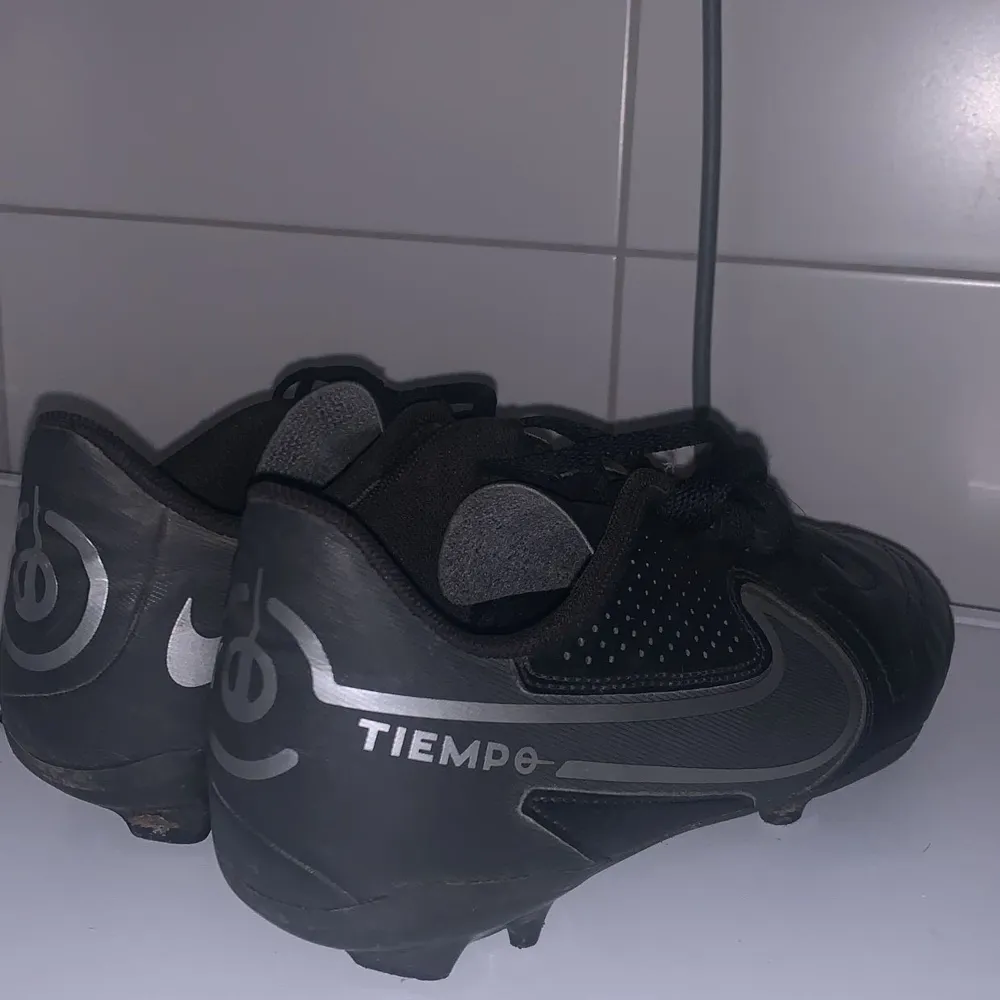 Säljer ett par svarta Nike Tiempo fotbollsskor i bra skick. De har en klassisk design med snörning och är perfekta för fotbollsträning eller match. Skorna är gjorda i skinn och har en stilren svart färg med silverdetaljer. Perfekta för dig som vill ha både stil och funktion på planen!. Skor.