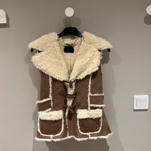 Brun mockaväst med foder från Vero Moda - Säljer en superfin brun mockaväst från Vero Moda i storlek M. Västen har ett mysigt vitt foder och snygga knytband framtill. Perfekt för höst och vinter, och den ger en härlig boho-vibe. Den har också praktiska fickor med foderkant. Ett måste för garderoben!