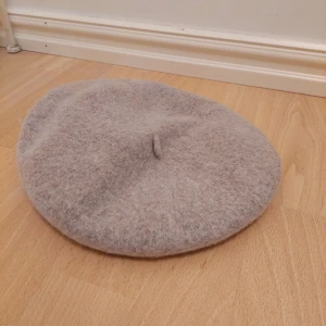Beige ullbasker - Säljer en superfin beige basker i ull. Perfekt för höst och vinter, den är både stilren och varm. Baskern har en klassisk design med en liten topp i mitten. Passar till alla outfits och ger en chic touch!