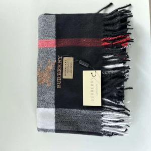  halsduk från Burberry - Säljer en superfin rutig halsduk från Burberry i 100% kashmir. Den har en klassisk design med svarta, gråa och röda rutor. Perfekt för att hålla sig varm och stilren under kyliga dagar. Den är i nytt skick och har fransar i ändarna. Passar perfekt för höst och vinter!