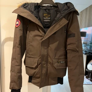 Mörk grön dunjacka från Canada Goose - Säljer en mörk grön dunjacka från Canada Goose i bra skick. Jackan har en fluffig pälsfodrad huva och praktiska fickor. Perfekt för kalla vinterdagar. Den har en dragkedja och muddar vid ärmsluten för extra värme. Passar perfekt för vinterns alla äventyr! 🧥❄️ små slit på insidan av ärmarna dock ej synligt när den är på