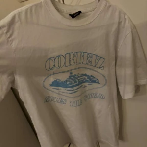 Corteiz T-shirt - Säljer en vit t-shirt från Cortez i storlek medium. Den har ett coolt blått tryck med texten 'Cortez Rules the World' och en bild av ett skepp. Perfekt för en avslappnad stil!