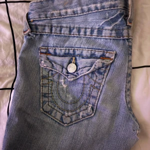 Lågmidjade bootcut jeans från True Religion - Säljer ett par snygga blå jeans från True Religion i bra skick. De har en klassisk femficksdesign med coola detaljer på bakfickorna och en knappgylf. Perfekta för vardagsbruk eller en avslappnad utekväll. Passar dig som gillar en stilren och tidlös look. En enstaka lagning som inte syns på utsidan, därav priset.❤️ fråga gärna 