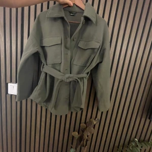 Militärgrön jacka med knytbälte - Säljer en snygg olivgrön jacka från GinaTricot i mycket bra skick. Den har en normal passform med långa ärmar och ett praktiskt knytbälte i midjan. Perfekt för höst och vår! Jackan har knappar framtill och två stora fickor på bröstet. Superchic och lätt att matcha med olika outfits. Nypris: 600kr