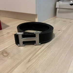 Svart skärp med silverfärgat spänne - Säljer ett stilrent svart skärp i skinn med ett stort silverfärgat H-spänne. Perfekt för att ge en lyxig touch till din outfit. Skärpet är i nytt skick och kommer i originalförpackning. Passar både till vardags och fest!