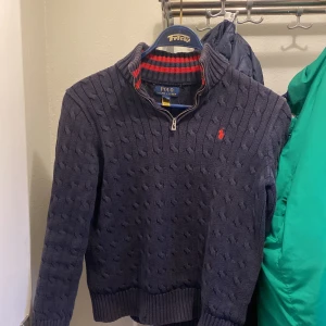 Mörkblå stickad tröja från Polo Ralph Lauren - Säljer en snygg mörkblå stickad tröja från Polo Ralph Lauren. Den har en dragkedja vid halsen och ett rött broderat märke på bröstet. Perfekt för kyliga dagar med sin ribbade krage och muddar. Passar både till vardags och lite finare tillfällen.