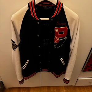 Varsitykofta från Polo Ralph Lauren - Säljer en snygg varsitykofta från Polo Ralph Lauren i blått och vitt. Jackan har broderade detaljer och knappar framtill. Perfekt för höst och vår med sin sportiga stil och långa ärmar. Den har coola patches och text på ryggen som ger en unik look.