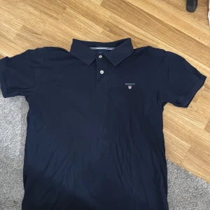 Mörkblå pikétröja från GANT - Säljer en snygg mörkblå pikétröja från GANT i storlek M. Tröjan har en klassisk krage med två knappar och GANT-loggan broderad på bröstet. Perfekt för en stilren och avslappnad look. Passar både till vardags och lite finare tillfällen. Sparsamt använd. 