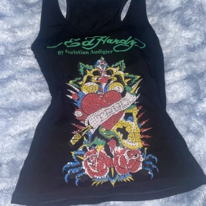 Svart linne från Ed Hardy - Coolt svart linne från Ed Hardy med ett färgglatt tryck av ett hjärta och rosor. Detaljerna är prydda med glittriga stenar som ger en extra touch, men några fåtal diamanter är borta. Perfekt för sommaren. Linnet är i bra skick och har en normal passform.
