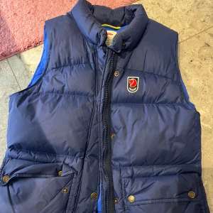 Blå dunväst från Fjällräven - Säljer en snygg blå dunväst från Fjällräven i damstorlek M. Västen har en dragkedja framtill och praktiska fickor med knappar. Perfekt för höst och vår när du vill hålla dig varm och stilren. 🦊