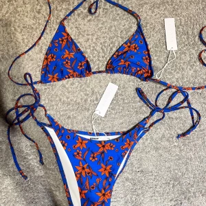 Blå blommig bikini med knytband - Säljer en supercool blå bikini från Bright Swimwear med orangea blommor. Den har knytband både vid nacken och sidorna av trosorna för snygg passform. ALDRIG ANVÄND MED ALLA LAPPAR OCH HYGIENSKYDD KVAR!! Nypris 750kr