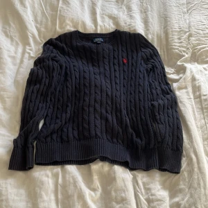 Marinblå Ralph lauren stickad tröja - Säljer en snygg svart stickad tröja från Polo Ralph Lauren. Den har ett klassiskt flätmönster och en liten röd logga på bröstet. Perfekt för kyliga dagar och passar både till vardags och lite finare tillfällen. Tröjan är i mycket bra skick och supermjuk!