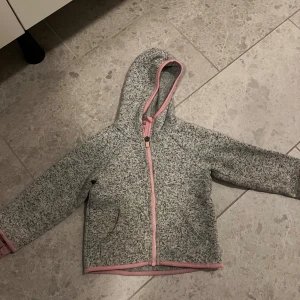 Grå fleecejacka från H&M - Mysig grå fleecejacka från H&M med rosa detaljer och dragkedja. Perfekt för kyliga dagar med sin mjuka och varma känsla. Jackan har en huva och långa ärmar, vilket gör den idealisk för höst och vår. Passar barn i storlek 110/116.