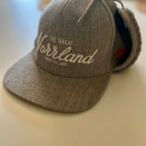 Grå keps med öronlappar från Sqrtn Company - Säljer en snygg grå keps med öronlappar från North Company. Den har texten 'The Great Norrland' broderad framtill och en liten orange blomma på sidan. Perfekt för kyliga dagar med sin mjuka insida. Passar både till vardags och utflykter! Nypris 500kr