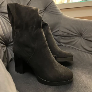 Svarta mockastövlar med klack - Snygga svarta knähöga stövlar i mocka med klack. Perfekta för höst och vinter, dessa boots har en stilren design som passar både till vardags och fest. De är i mycket bra skick och ger en elegant touch till vilken outfit som helst. Endast lite damm på skorna som lätt går att ta bort