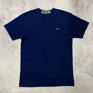 Mörkblå t-shirt från Comme des Garçons - Säljer en snygg mörkblå t-shirt från Comme des Garçons med det ikoniska hjärtmärket med ögon på bröstet. T-shirten är i bomull och har en normal passform. Perfekt för en stilren och avslappnad look!